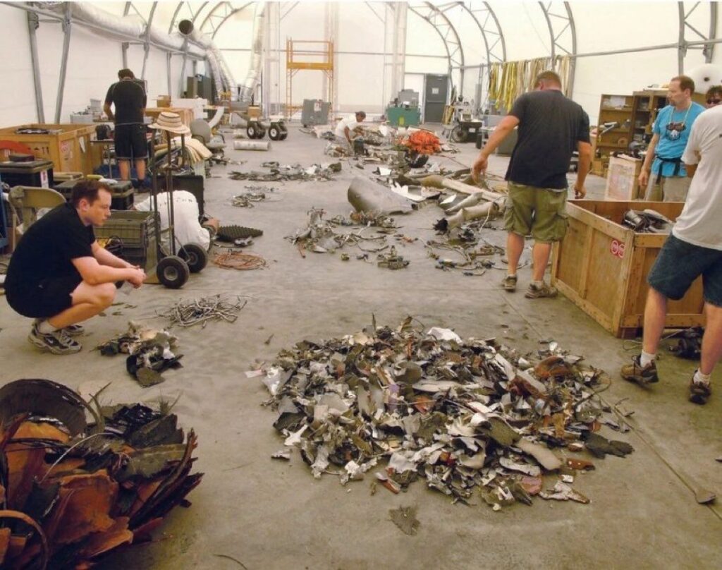 SpaceX IPO - Workshop with workers sorting piles of SpaceX Falcon 1 scrap metal on a concrete floor inside a large hangar-like space to one side and equipment around them.